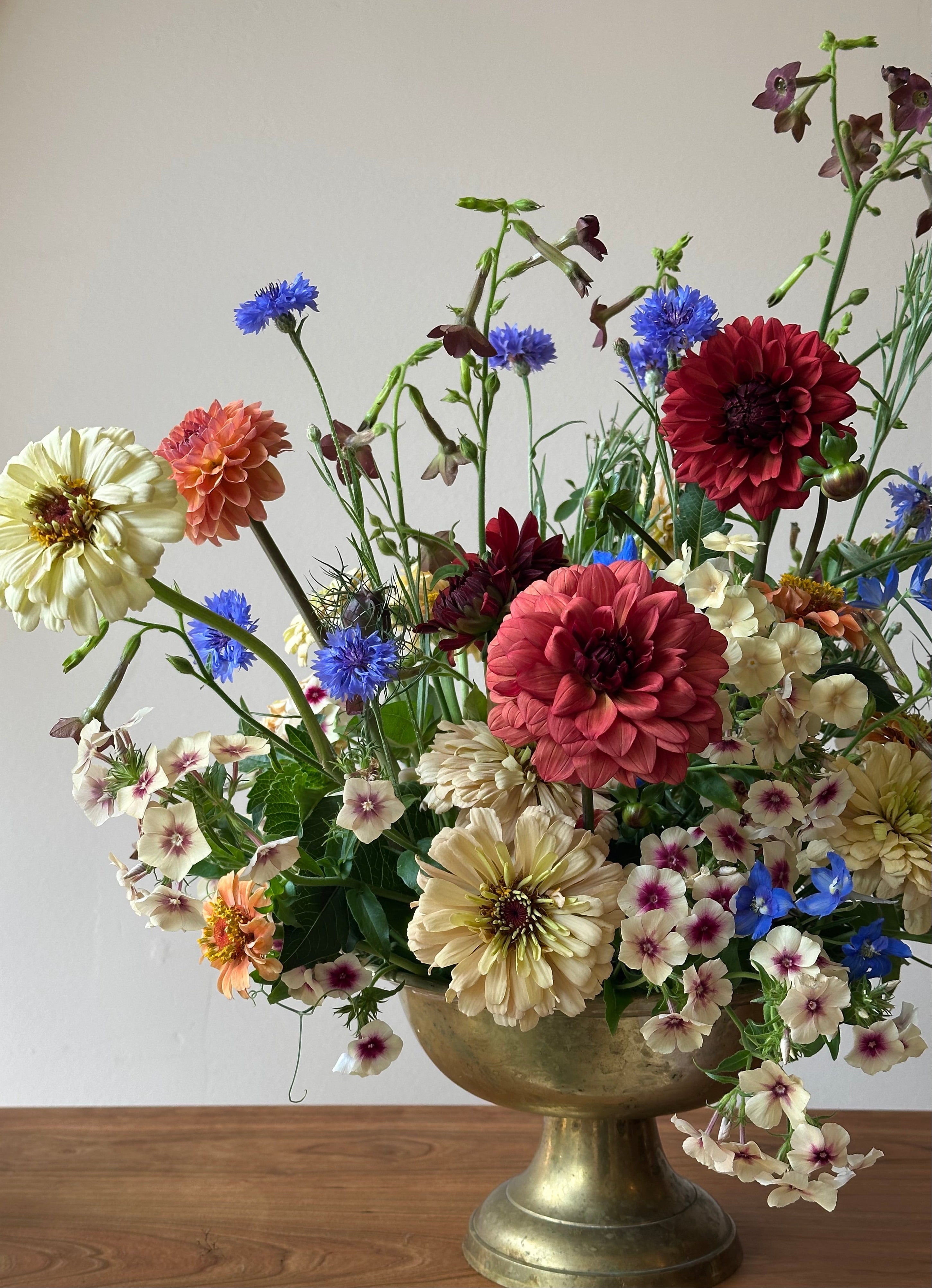 A vibrant, multicolored wildflower arrangement in a vintage-style bronze pedestal vase, set against a neutral beige wall and wooden tabletop.