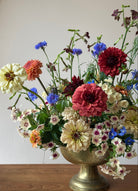 A vibrant, multicolored wildflower arrangement in a vintage-style bronze pedestal vase, set against a neutral beige wall and wooden tabletop.